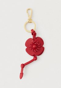 Sarkanas ādas ziedu piekariņš ar zelta metāla gredzenu un āķi, kuram ir mezglota siksniņa detaļa uz gaiša fona.