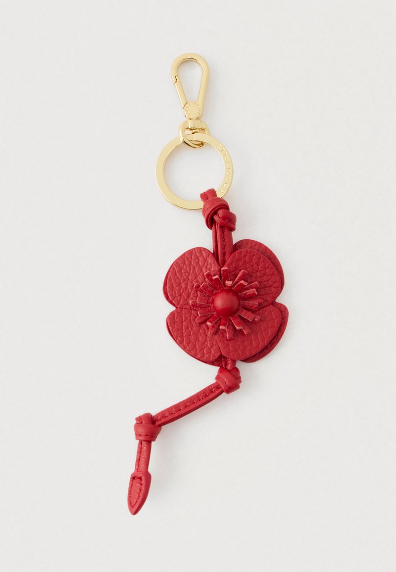 Sarkanas ādas ziedu piekariņš ar zelta metāla gredzenu un āķi, kuram ir mezglota siksniņa detaļa uz gaiša fona.