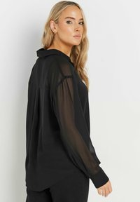Camicia nera trasparente con colletto, maniche lunghe trasparenti e orlo leggermente curvo. Il tessuto è leggero e con una texture liscia.