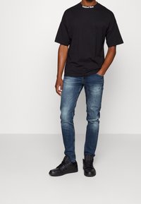 Camiseta negra de manga corta con el texto "Refined Rebel" en el cuello, combinada con jeans slim-fit de un azul medio y zapatillas negras.