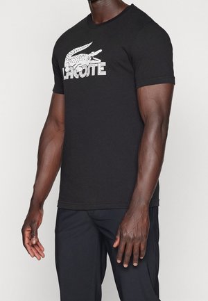 Homme portant un t-shirt noir Lacoste avec un logo crocodile blanc et un pantalon noir assorti sur un fond uni.