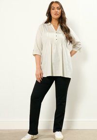 Hellbeige Tunika mit Kragen, Falten und Knopfdetails, kombiniert mit schwarzen geraden Hosen und weißen Sneakers. Glatte Stoffoberfläche.