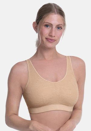 Frau mit blond zurückgebundenem Haar, die einen beigen Sport-BH mit Spitzenmuster trägt und sanft vor einem schlichten hellen Hintergrund lächelt.