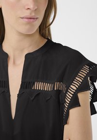 Detailní záběr ženy nosící černou halenku s výřezy na ramenou a horní části hrudníku, s výstřihem do V.