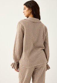 Nainen, jolla on yllään ruskea ja valkoinen ruudullinen pitkähihaisen paita ja siihen sopivat housut, katsoo sivulle yksinkertaista vaaleaa taustaa vasten.