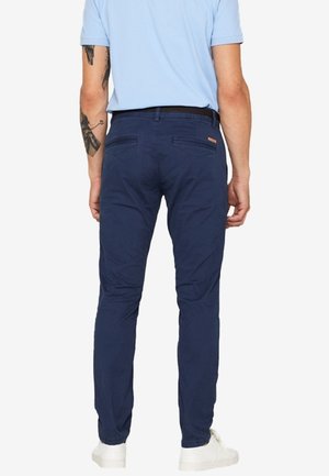 Homme portant un pantalon bleu marine, une chemise à manches courtes bleu clair, des baskets blanches et des tatouages visibles sur le bras gauche, debout de dos.