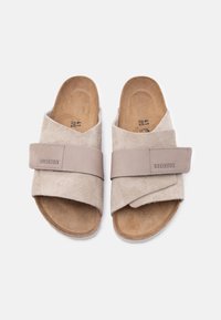 Ζευγάρι μπεζ σανδαλιών Birkenstock slip-on με ρυθμιζόμενους ιμάντες και σουέτ εσωτερική σόλα σε λευκό φόντο.
