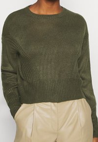 Olijfgroen gebreid sweater met een geribbelde textuur, ronde halslijn en een cropped pasvorm, gecombineerd met beige leren broek.
