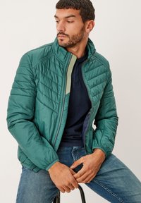 Giacca trapuntata verde con colletto alto e chiusura con zip, con polsini elastici e una camicia blu sotto. Indossata con jeans blu.