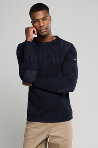 Granatowy sweter z prążkowanej dzianiny z długimi rękawami, z łatami na łokciach i okrągłym dekoltem. W połączeniu z jasnobeżowymi spodniami.