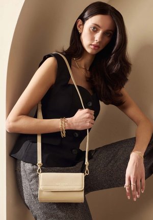 Sac bandoulière en crème texturé avec quincaillerie dorée, présentant une fermeture à rabat et une sangle réglable, associé à un haut noir sur mesure et un pantalon gris.