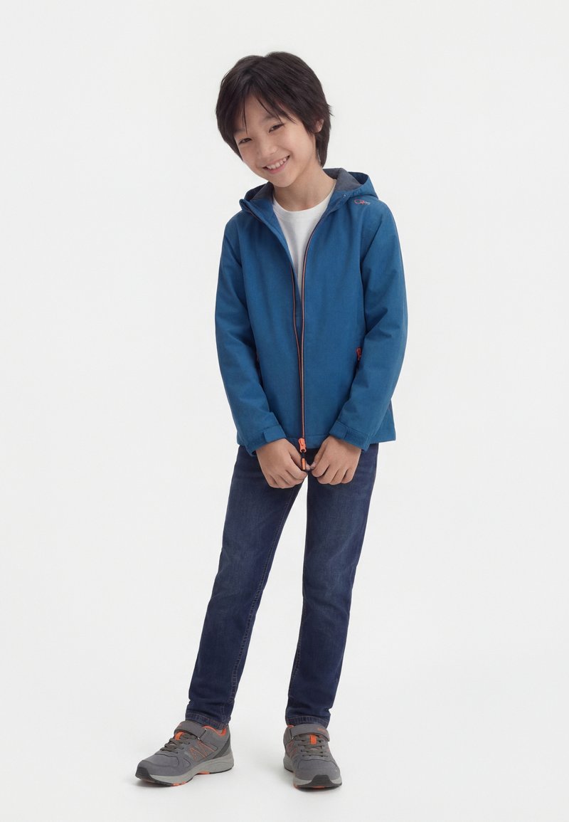 Χαμογελαστό παιδί που φοράει μπλε μπουφάν με κουκούλα, λευκό πουκάμισο, σκούρο τζιν και γκρι αθλητικά παπούτσια, στέκεται με τα χέρια σταυρωμένα μπροστά.