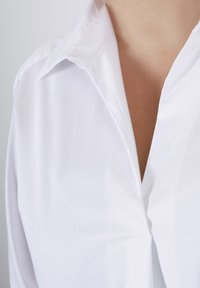 Witte blouse met een klassieke kraag, een gladde textuur en een diepe V-halslijn. De stof is lichtgewicht en valt zacht.