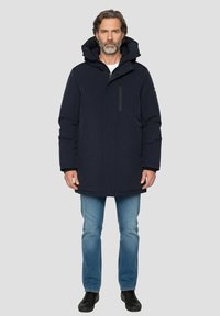 Donkerblauwe gewatteerde jas met capuchon, voorzien van een ritssluiting aan de voorkant en een borstzak. Gedragen over een wit shirt en een blauwe spijkerbroek. Zwarte schoenen.