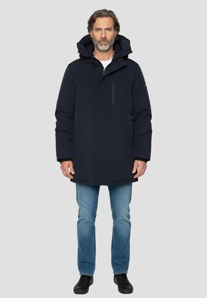 Donkerblauwe gewatteerde jas met capuchon, voorzien van een ritssluiting aan de voorkant en een borstzak. Gedragen over een wit shirt en een blauwe spijkerbroek. Zwarte schoenen.