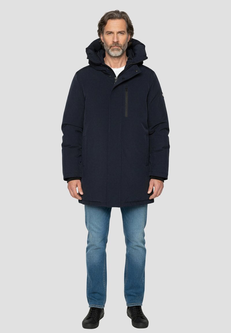 Donkerblauwe gewatteerde jas met capuchon, voorzien van een ritssluiting aan de voorkant en een borstzak. Gedragen over een wit shirt en een blauwe spijkerbroek. Zwarte schoenen.