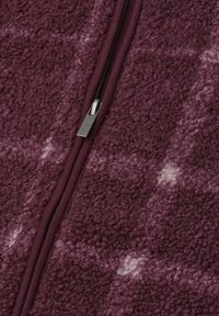 Gros plan d'un tissu polaire texturé bordeaux avec une fermeture éclair noire centrale et un curseur argenté.