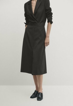 Femme portant une robe blazer noire à manches retroussées et des mocassins noirs, debout devant un fond blanc uni.