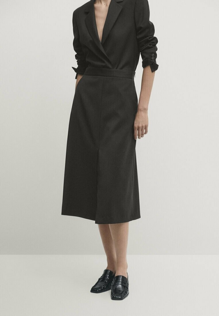 Femme portant une robe blazer noire à manches retroussées et des mocassins noirs, debout devant un fond blanc uni.
