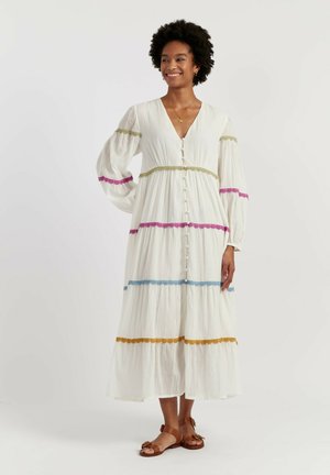 Donna sorridente con capelli ricci indossa un lungo vestito bianco abbottonato con fasce orizzontali di pizzo colorato e sandali marroni, in piedi su sfondo bianco.