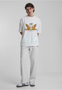Hvit oversized t-skjorte med grafisk trykk i oransje og svart, sammen med grå joggebukser. Svarte og hvite joggesko fullfører antrekket.