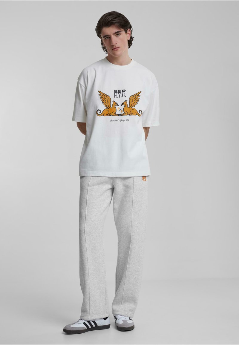 Hvit oversized t-skjorte med grafisk trykk i oransje og svart, sammen med grå joggebukser. Svarte og hvite joggesko fullfører antrekket.