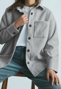 Light gray wool shirt jacket with a collared neck, button closure, single chest pocket, and slightly curved hem, worn over a white shirt.