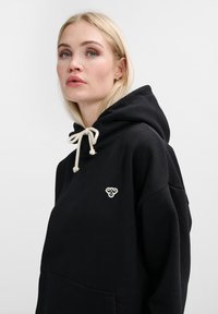 Svart huvtröja med en mjuk textur, utrustad med en dragsko med krämfärgade ändar och en liten broderad logo på bröstet.