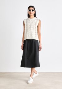 Pulover tricotat fără mâneci, de culoare albă murdară, cu un model texturat, asortat cu o fustă lungă neagră și adidași albi. Design simplu, modern.