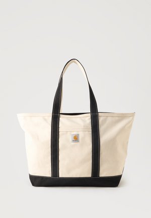 Beżowa torba typu tote z płótna z czarnymi uchwytami i spodem, przednią kieszenią oraz naszywką z logo Carhartt umieszczoną centralnie nad kieszenią.
