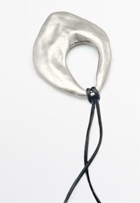 Ciondolo in argento con una forma organica e astratta, caratterizzato da un taglio centrale e una corda in pelle nera attaccata. Texture liscia, design minimalista.