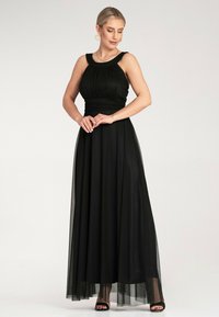 Czarna wieczorowa suknia z dopasowanym gorsetem i plisowanymi detalami, z przezroczystą warstwą i zwiewnym dołem. Podkreślona szerokim dekoltem.