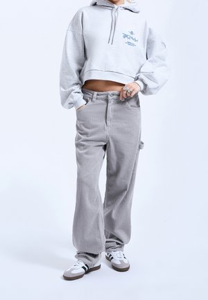 Osoba ubrana w krótką jasnoszarą bluzę z kapturem, szerokie spodnie z sztruksu w kolorze szarym oraz białe trampki z czarnymi paskami, stojąca z jedną ręką w kieszeni.