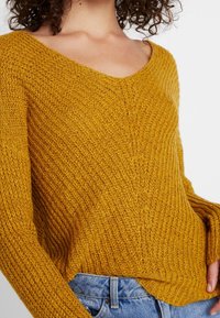 Hořčičně žlutý pletený svetr s výstřihem do V, ribbovanou texturou a dlouhými rukávy. Doplňuje ho mírně zakřivený lem.