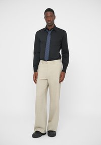 Schwarzes Langarmhemd mit knöpfbarem Kragen, grau gestreifte Krawatte, beige weite Hose und schwarze Slipper. Sauberer, maßgeschneiderter Look.