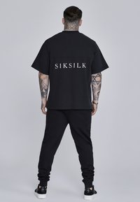 Melna lielā izmēra t-kreklu ar "SIKSILK" logo uz muguras, apvienots ar melnajām biksēm un melnajām kedām neitrālā fonā.