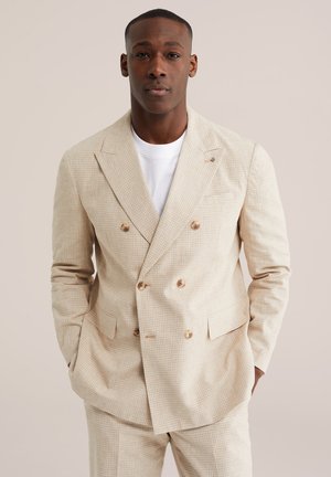 Un homme debout les mains dans les poches, portant un costume croisé beige et une chemise blanche, sur un fond neutre uni.