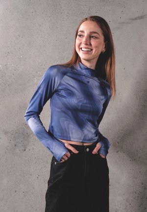 Mujer sonriente con el pelo largo lleva un top corto de manga larga azul con estampado y pantalones negros, de pie frente a una pared de hormigón gris.