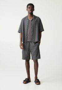 Grå kortærmet skjorte med knaplukning over en grå tanktop, parret med grå shorts og sorte sandaler, med en afslappet pasform og glat tekstur.