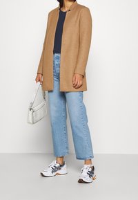 Brun skræddersyet frakke over en marineblå top, parret med lyseblå jeans med lige ben og hvide sneakers med sorte detaljer. Hvid tekstureret håndtaske.