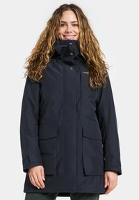 Young woman with long curly hair wearing a dark navy hooded raincoat with front pockets and high collar, smiling against a light background.