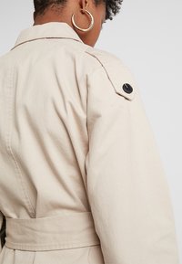 Trench coat beige con vita regolabile, caratterizzato da un colletto a punta, dettagli con bottoni neri sulle maniche e un tessuto liscio e testurizzato.