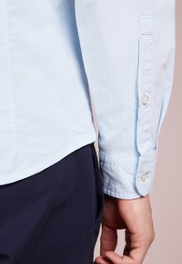 Světle modrá košile s dlouhým rukávem a hladkou texturou, s manžetou na knoflíky a zaobleným lemem. Kombinováno s tmavě námořnickými kalhotami.