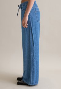 Pantalons en denim bleu clair à jambes larges, avec une taille à cordon de serrage, des poches latérales et une coupe décontractée, présentant une finition texturée et des bords ourlés.