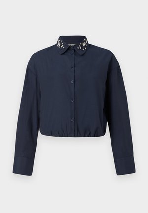 Mornarščina modra skrajšana majica z ovratnikom na gumb, ki vsebuje okrasne dodatke na ovratniku. Ima dolge rokave in elastiko na robu.