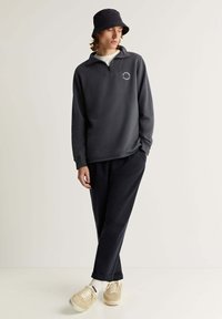 Grijze fleece pullover met een kwartrits, voorzien van een logo. Gecombineerd met donkere broeken en beige sneakers, aangevuld met een zwarte bucket hat.