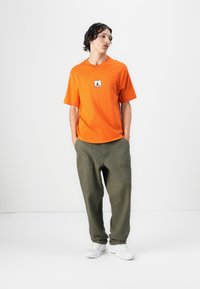 Orange t-shirt med et logo patch, parret med olivengrønne, afslappede bukser og hvide sneakers, der viser et afslappet outfit.