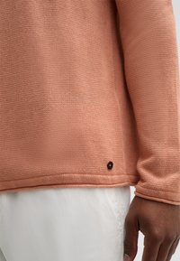 Maglione lavorato a maglia rosa chiaro con maniche lunghe, scollo rotondo e un piccolo logo nero sull'orlo, indossato sopra pantaloni bianchi.