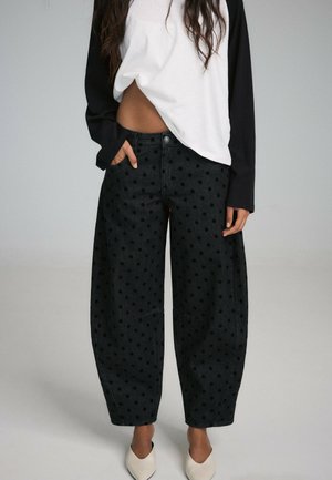 Femme portant un pantalon large noir à pois, des chaussures blanches pointues et un haut à manches longues blanc et noir avec une main dans la poche.