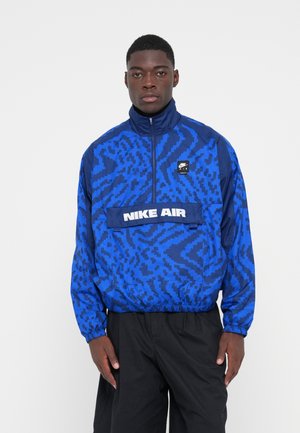 Nike Air windbreaker jack in blauw met een geometrisch patroon, kwart-rits ontwerp, elastische manchetten en een logo patch aan de voorkant.